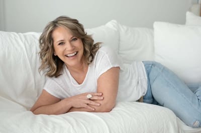 Lisa laughing as she leans on white linen cushions, wearing a white t-shirt and jeans.