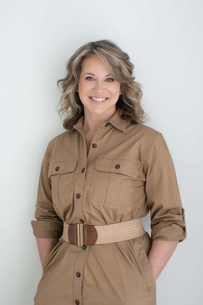 Lisa smiling in a khaki shirt dress with a woven belt, hands in her pockets.