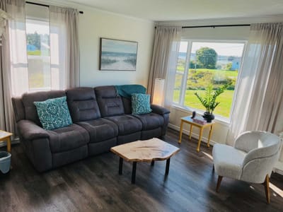 The counselling room: a deep grey sofa with teal cushions, a live-edge wooden coffee table, a soft chair, and a large window looking over green fields toward the water.