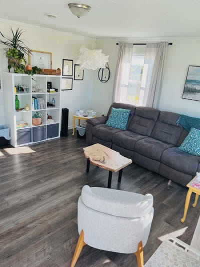 The counselling room: a white bookshelf with books, plants, essential oils and a salt lamp, framed certificates on the wall, and a grey sofa with teal cushions.